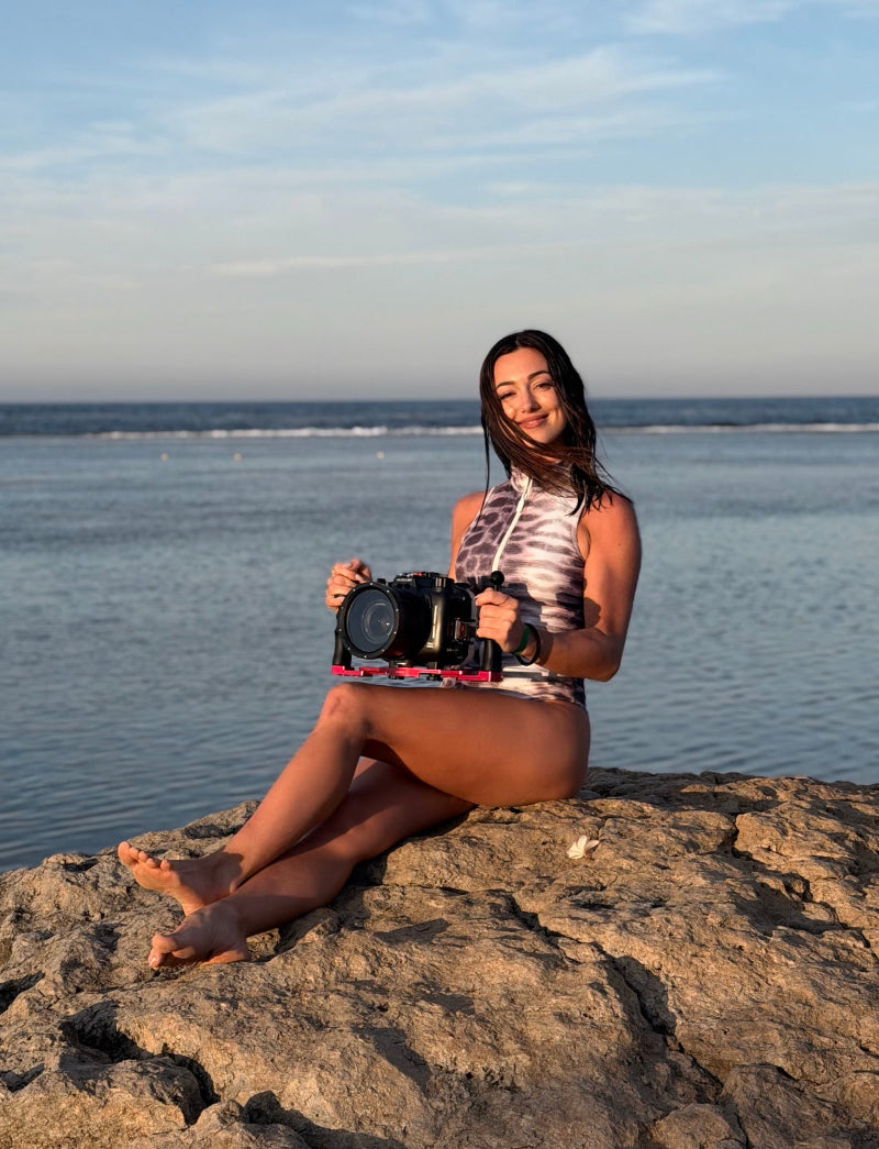 A woman sitting on a rocky shoreline by the ocean, holding a black camera mounted on a red and black rig. She is wearing a sleeveless patterned top and shorts, with the sea and sky in the background.