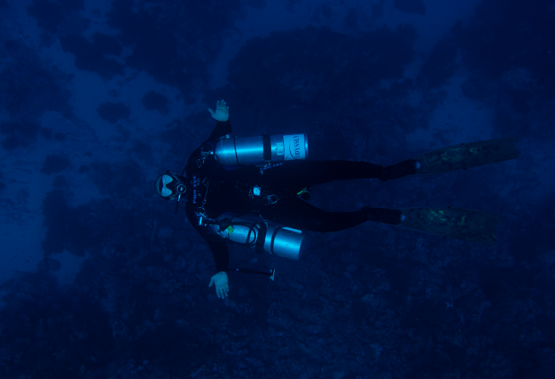 An underwater shot of a scuba diver swimming horizontally against a dark blue ocean background.
