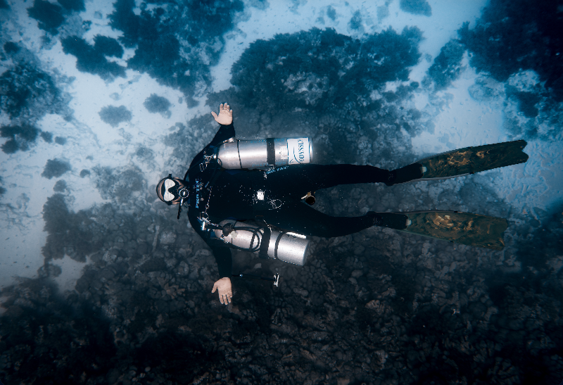 A scuba diver floating underwater above a dark coral reef, wearing a black wetsuit, regulator, and fins, with blue water and light filtering through from above.