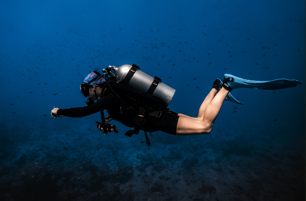 A scuba diver swims horizontally underwater in deep blue water, wearing a black wetsuit, silver oxygen tank, and blue fins.